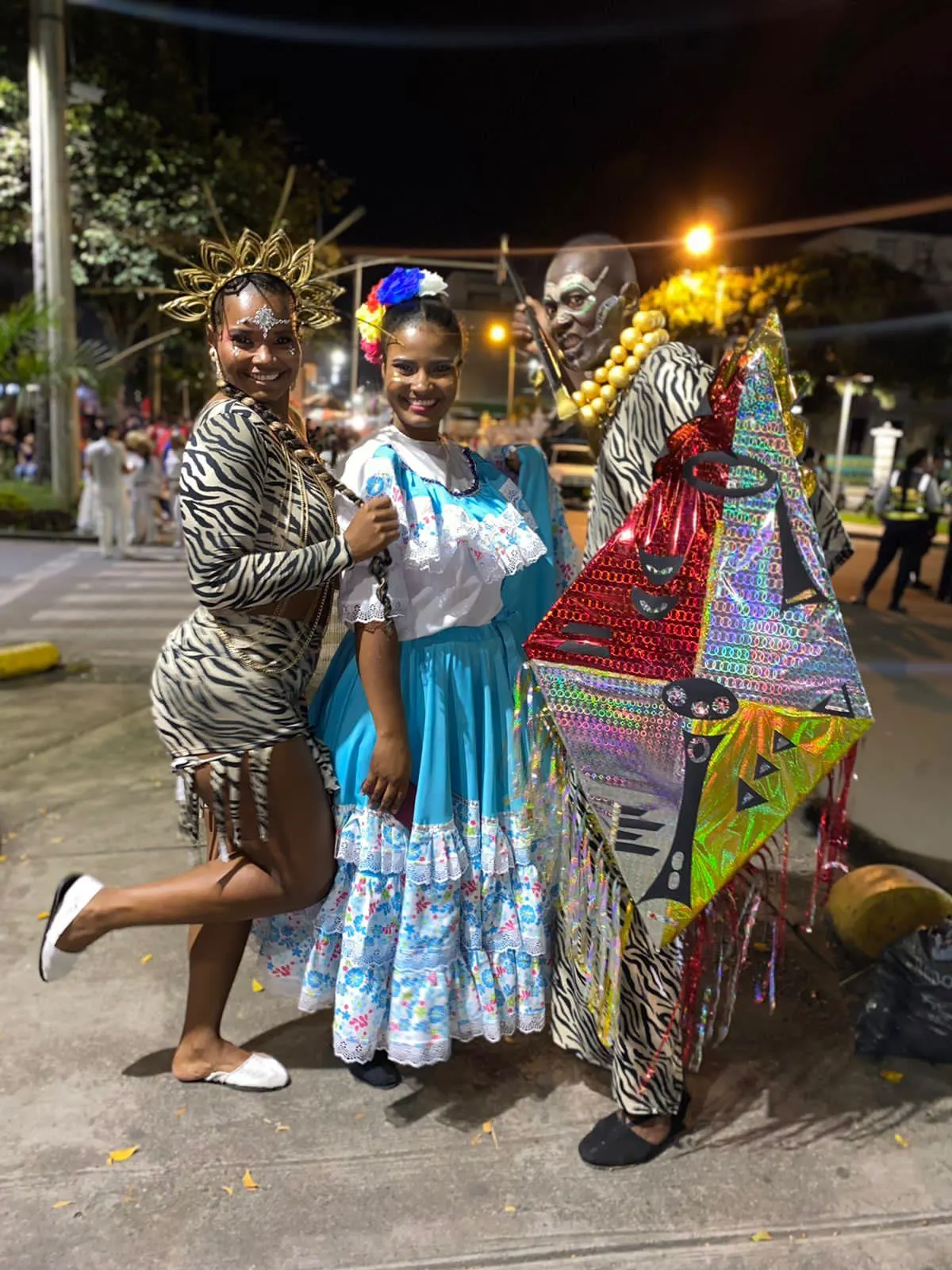 Tres integrantes de comparsa con vestuarios festivos, tocado dorado y cometa decorada en papel metalizado durante un desfile nocturno.