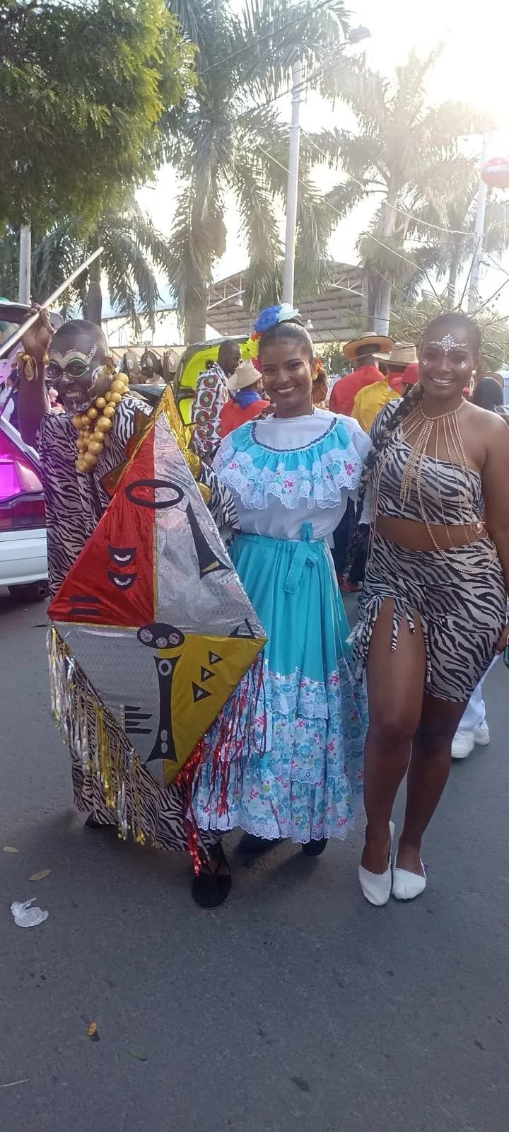 Comparsa diurna en la calle con traje folclórico azul celeste y figura enmascarada que porta una cometa artesanal decorada.