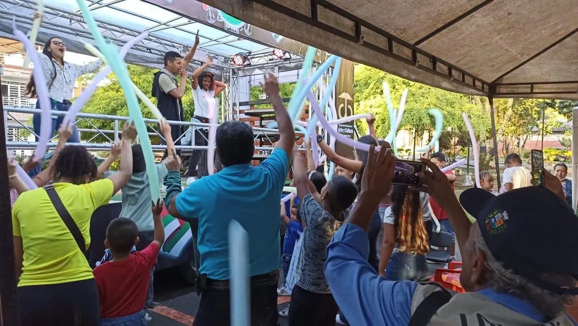 Público participando con globos alargados frente a una tarima del programa Arte y Cultura en un evento comunitario.