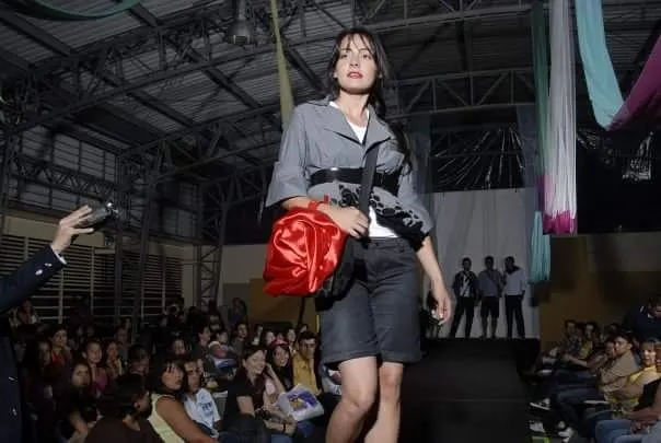 Modelo desfila en pasarela con chaqueta gris, fajín negro y bolso rojo de satén, vestuario de Daniel Cadavid, ante el público.