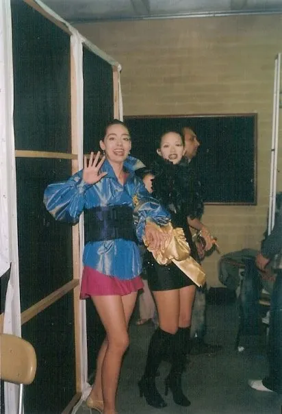 Dos modelos tras bambalinas con vestuarios de la colección: blusa azul de tafetán con fajín y minifalda fucsia, y conjunto negro con boa de plumas.