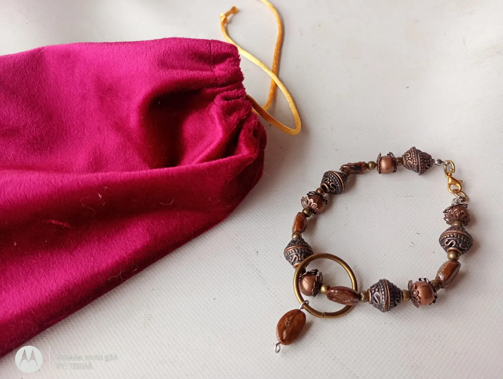 Pulsera con cuentas cobrizas y aro colgante junto a una bolsa de terciopelo fucsia, empaque artesanal de TEKNA.