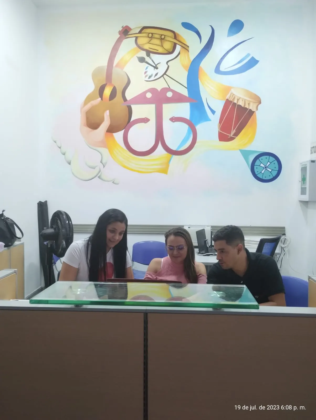 Tres integrantes del equipo trabajan frente a un computador bajo un mural con guitarra, tambora, paleta de pintura y símbolos culturales.