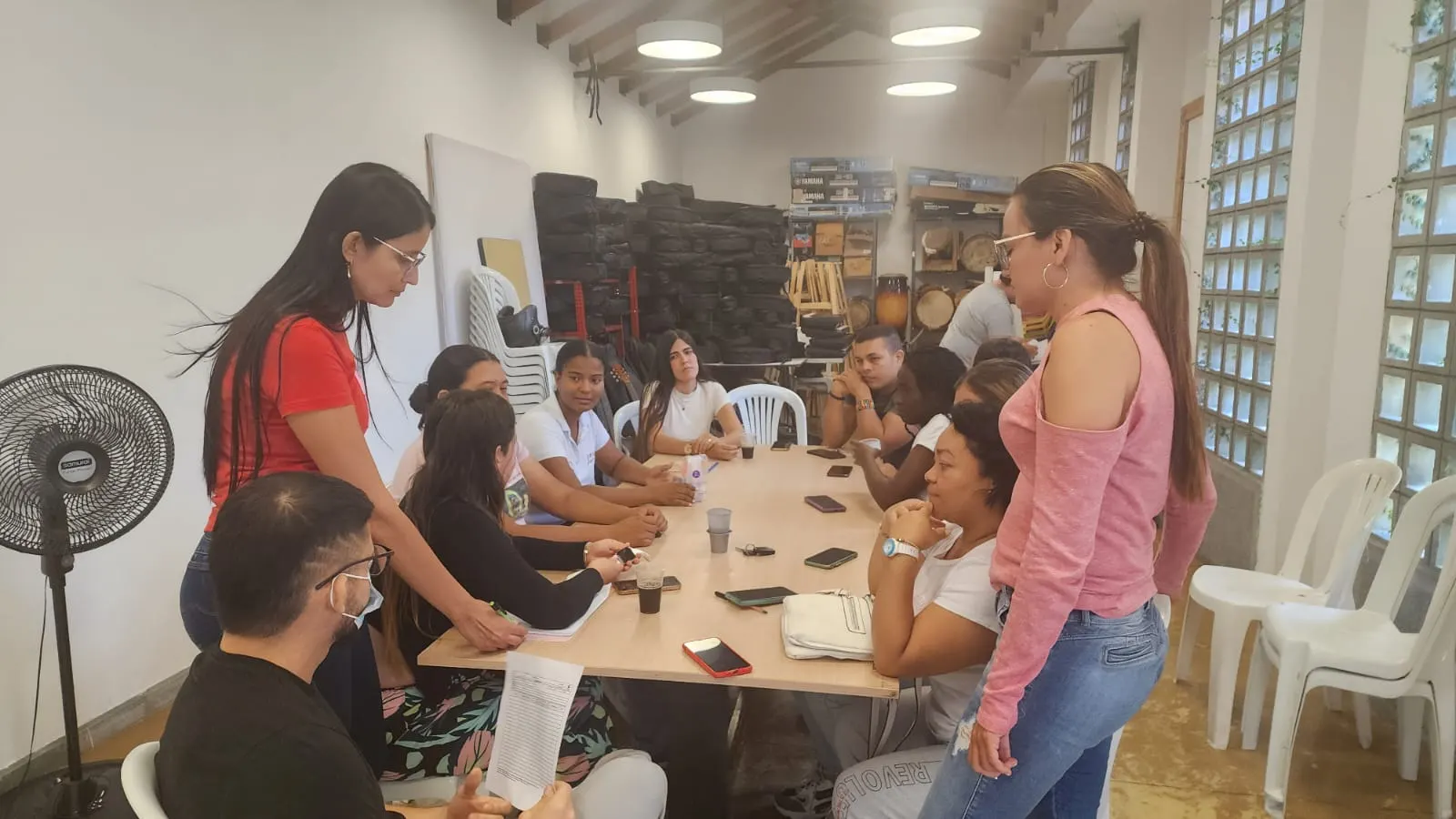 Reunión comunitaria en un salón cultural con instrumentos musicales almacenados al fondo; los participantes conversan alrededor de una mesa larga.