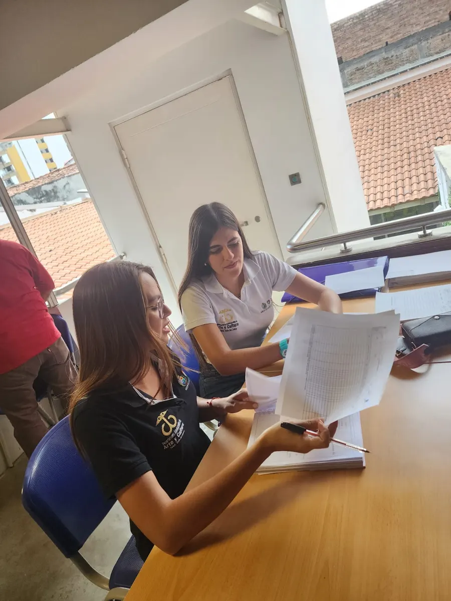 Gestora con camiseta del Departamento de Arte y Cultura de Tuluá revisa documentos de un proyecto junto a una compañera, en un balcón con tejados al fondo.