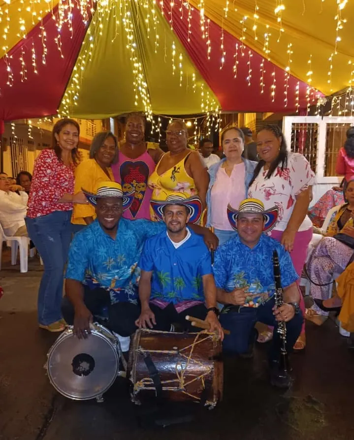 Trío de músicos con sombreros vueltiaos, tambora, redoblante y clarinete posa con integrantes de la comunidad bajo un toldo festivo iluminado.