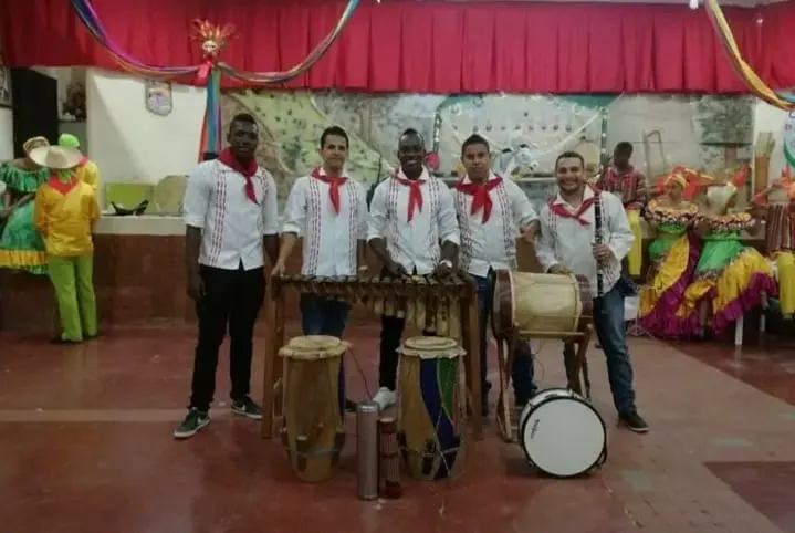 Agrupación de chirimía con marimba, tamboras, redoblante y clarinete sobre un escenario con telón rojo y trajes folclóricos al fondo.