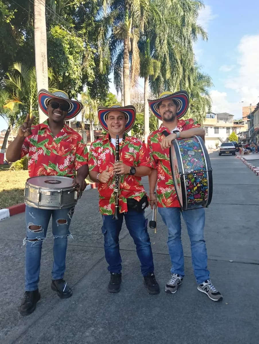 Franko Piñerez, al centro con clarinete y sombrero vueltiao, junto a dos percusionistas de su banda papayera con camisas florales en una calle arbolada.