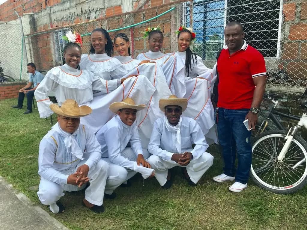 Agrupación de danza folclórica con trajes blancos tradicionales y tocados de flores; Jesús Albeiro Angulo acompaña al grupo antes de la presentación.