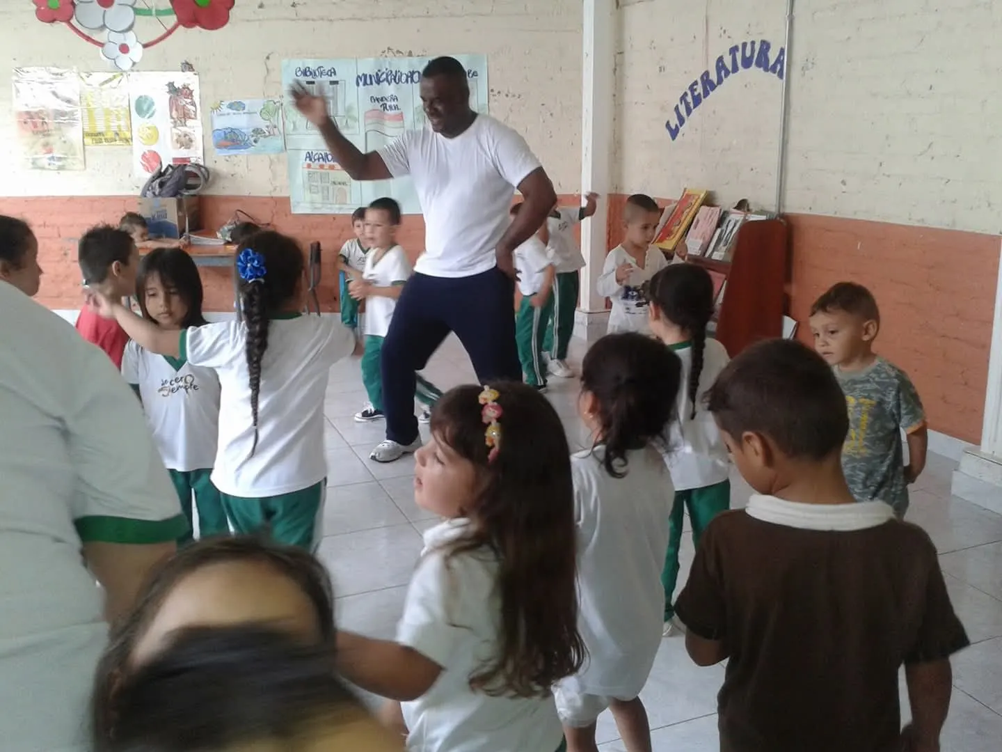 Sesión de danza con niñas y niños de primera infancia en un aula escolar; el formador guía los movimientos del grupo.