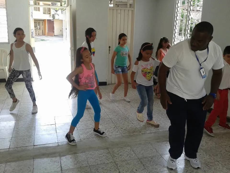 Clase de danza con niñas y adolescentes en un salón comunitario; el formador demuestra los pasos frente al grupo.