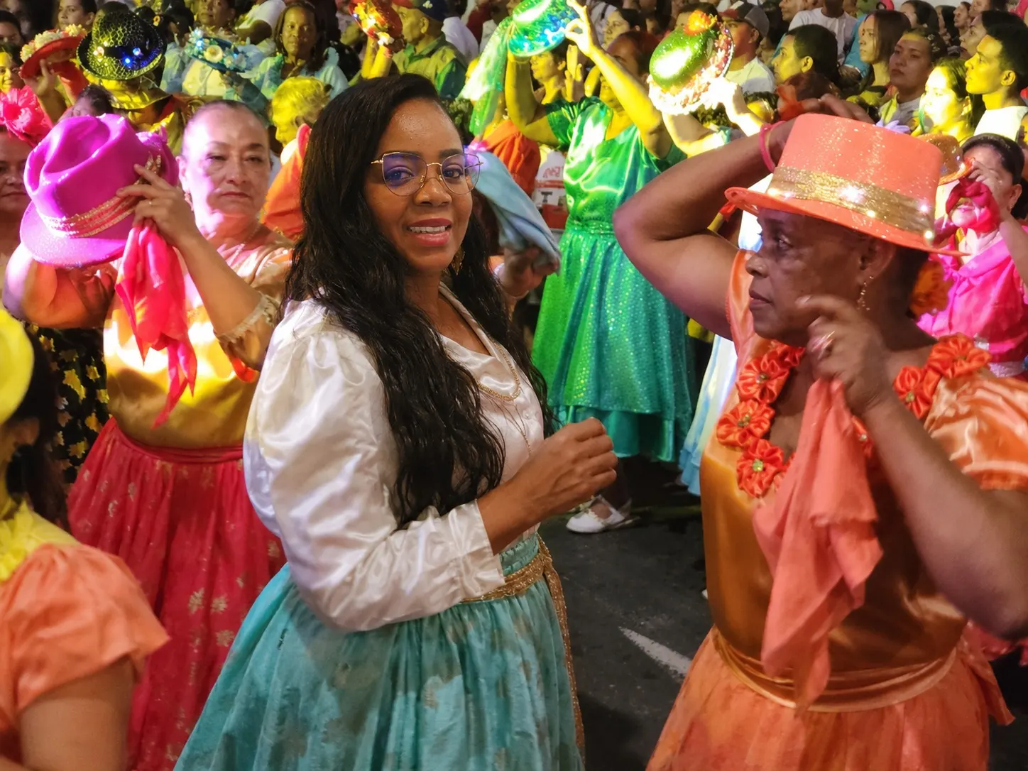 Martha participa en una danza folclórica nocturna con blusa blanca y falda verde aguamarina, rodeada de bailarinas con sombreros y vestidos de colores vivos.