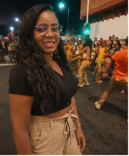 Martha Isabel Torres Vidal sonríe en una calle durante un desfile nocturno, con una comparsa de bailarines de vestuario amarillo al fondo.