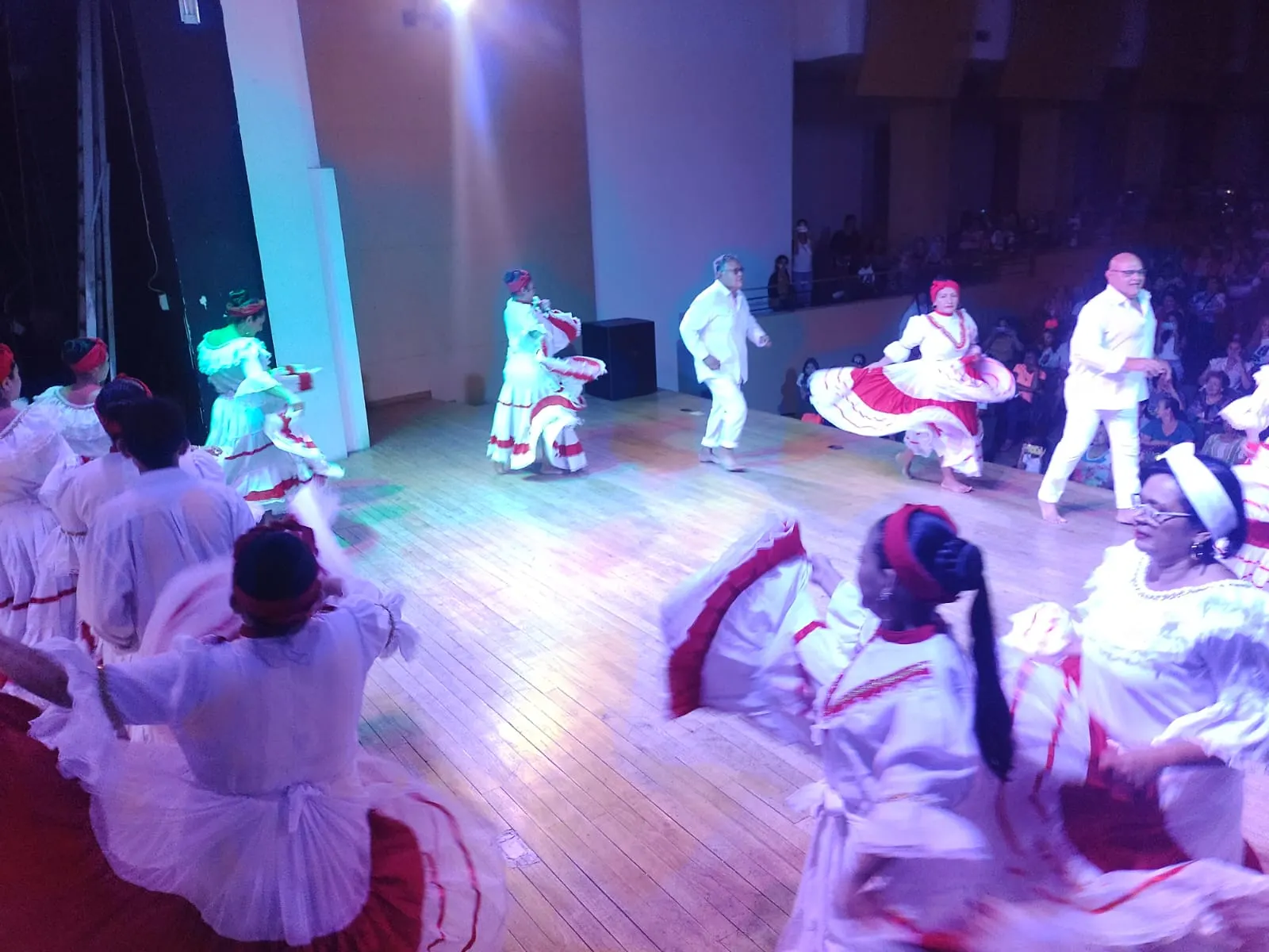 Presentación de danza folclórica en un teatro: parejas con trajes blancos y faldas rojas y blancas giran sobre el escenario ante el público.