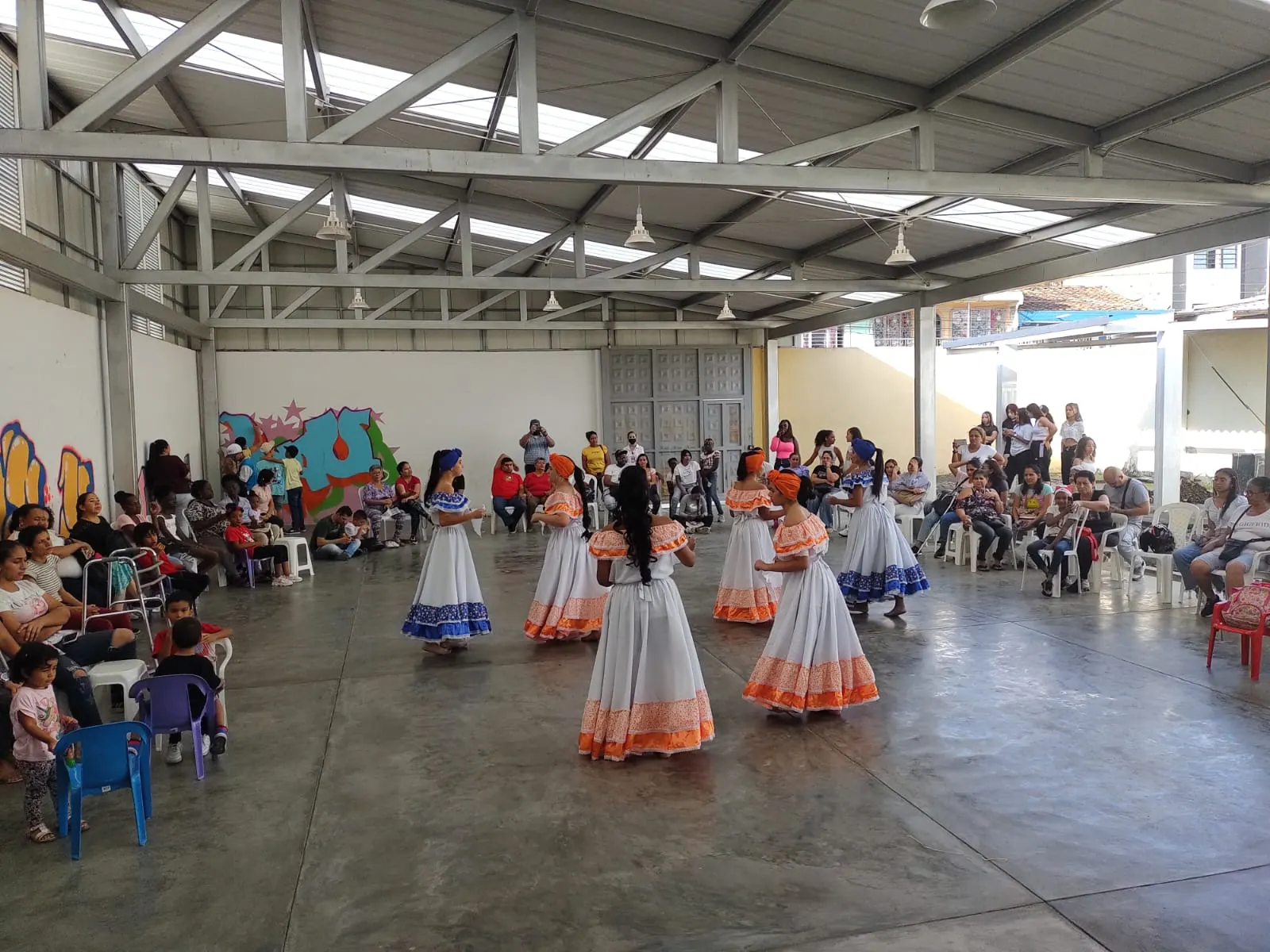 Muestra de danza folclórica en un espacio comunitario: bailarinas con turbantes y faldas amplias en blanco, azul y naranja danzan en círculo ante los asistentes.
