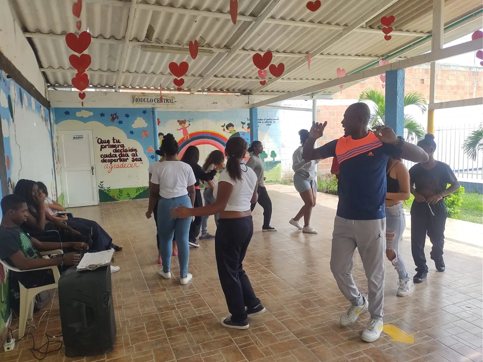 Orlando guía un ejercicio de baile con adolescentes en un patio cubierto decorado con corazones de papel.