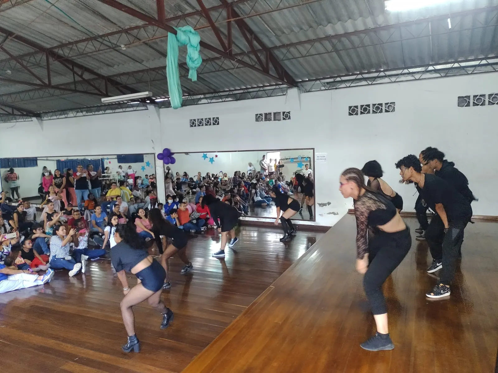 Agrupación juvenil de danza se presenta sobre una tarima de madera ante un público numeroso en un salón con espejos.