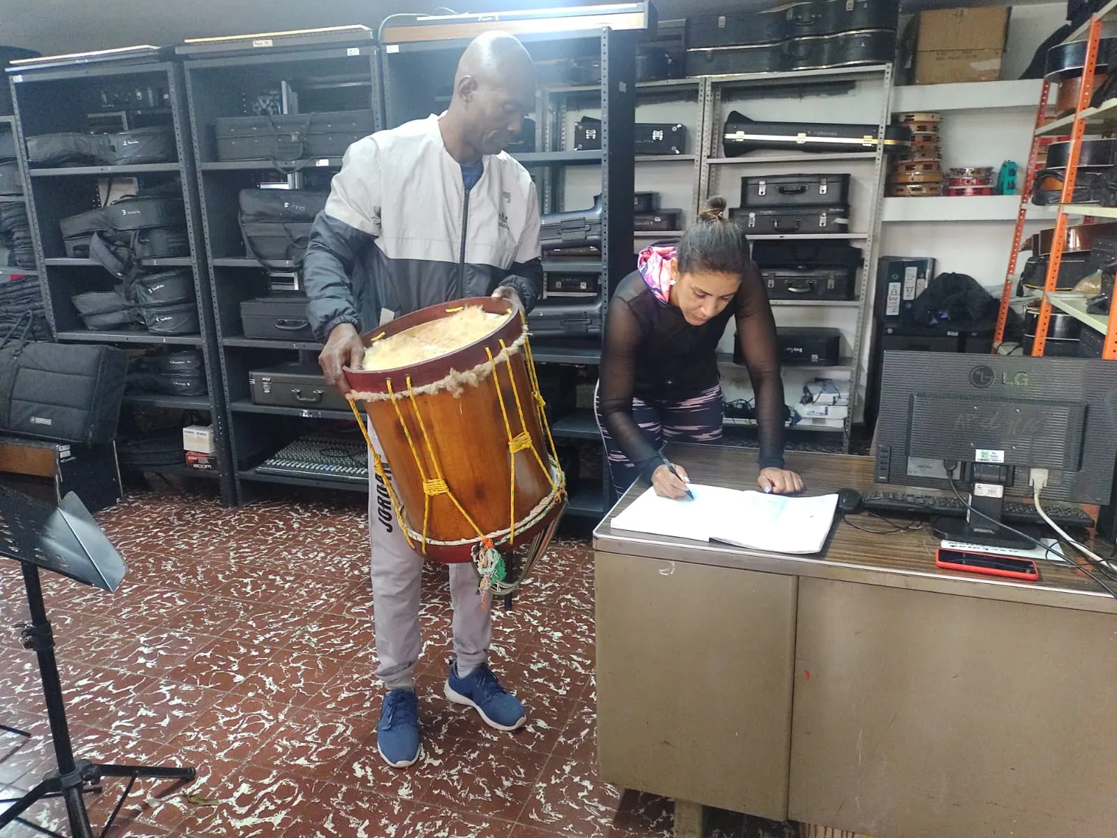 Orlando sostiene una tambora tradicional en un depósito de instrumentos musicales mientras una compañera diligencia un registro.
