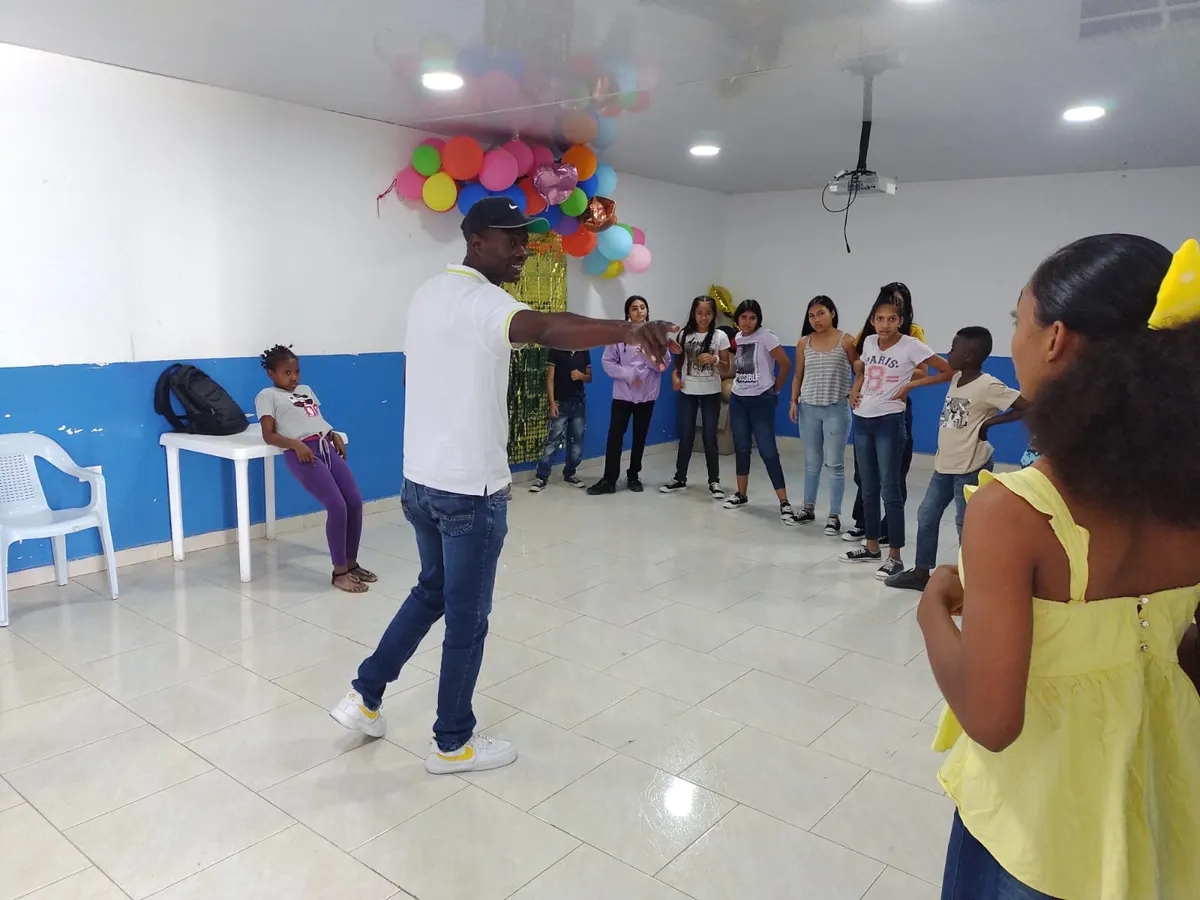 Orlando Solís Caicedo, con gorra y polo blanco, dirige un taller de danza ante un grupo de niños y jóvenes en un salón comunitario decorado con globos.