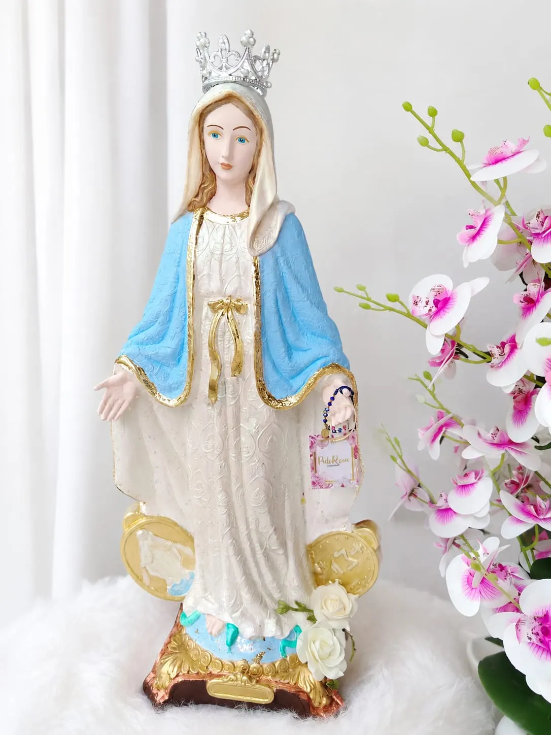 Imagen religiosa de la Virgen decorada en tonos azul, blanco y dorado, presentada junto a orquídeas con etiqueta de Palo Rosa Decoración.