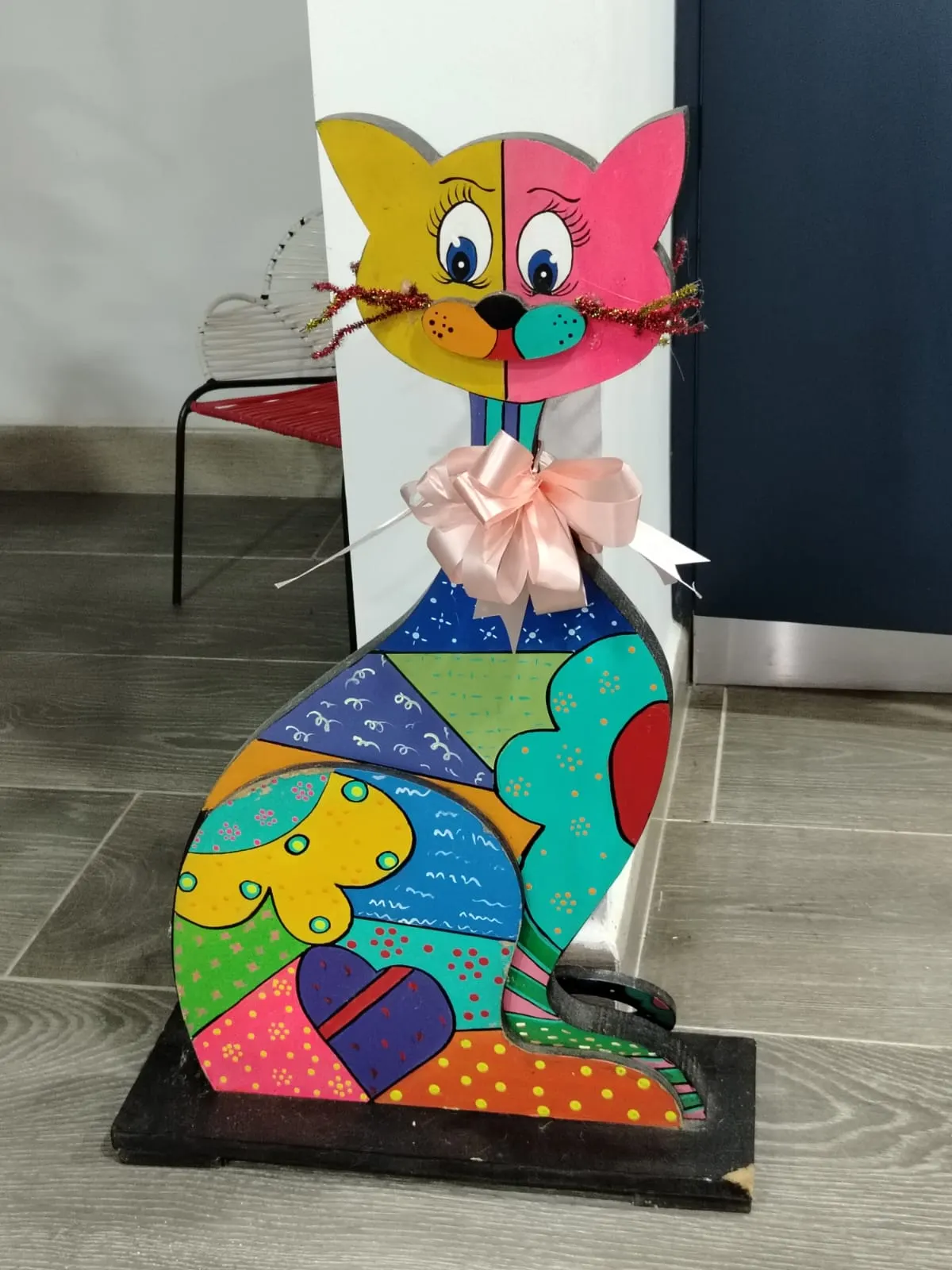 Gato decorativo de madera pintado a mano con patrones geométricos multicolores tipo pop art y lazo rosado al cuello.