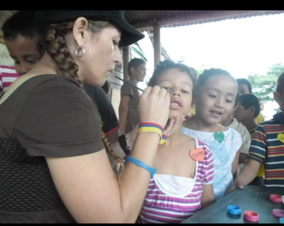 Sandra Milena Restrepo, con gorra y trenza, pinta el rostro de una niña durante un taller comunitario de pintacaritas rodeada de niños.