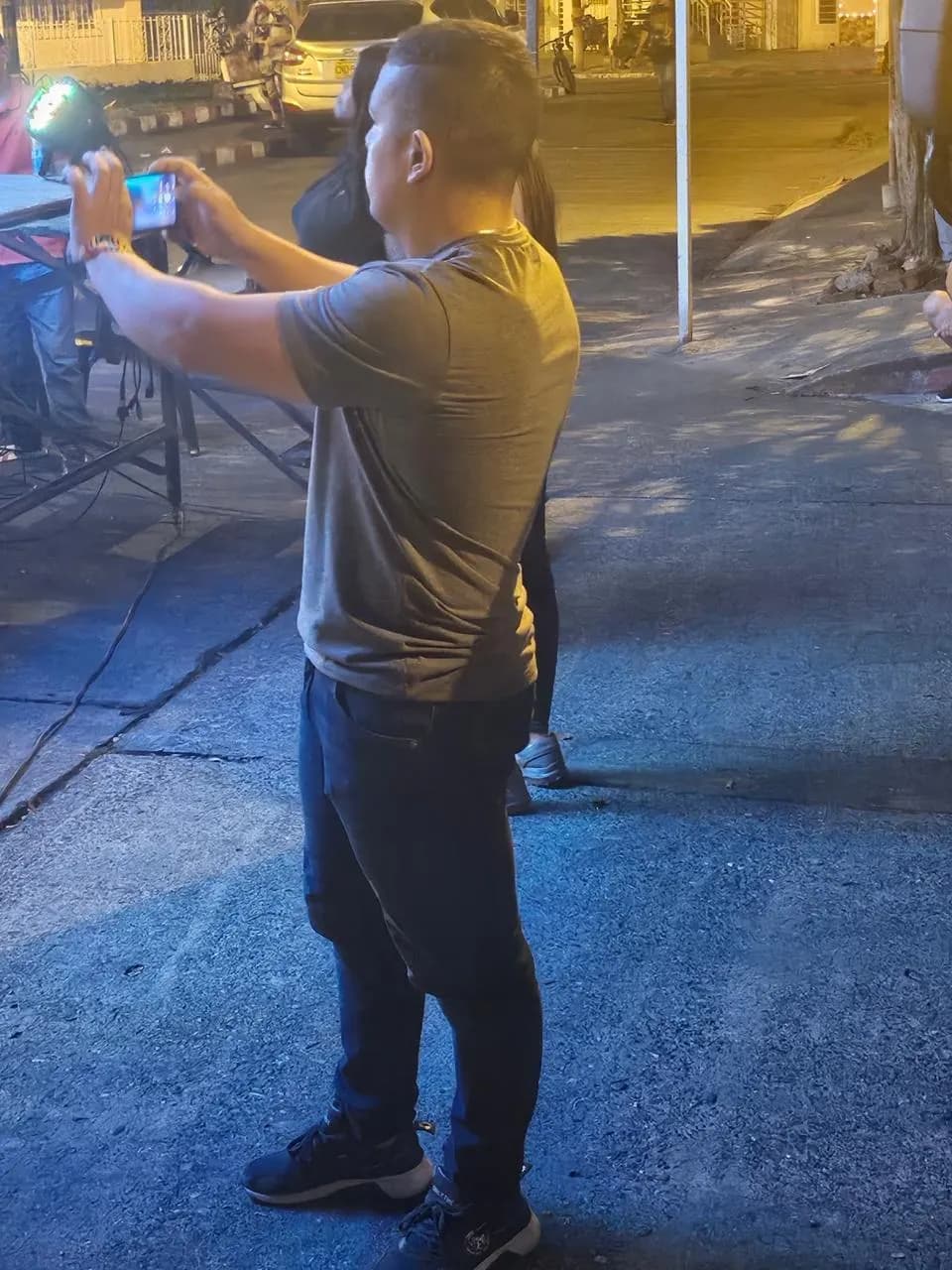 Registro nocturno de trabajo en campo: el gestor documenta con su celular una actividad cultural en el espacio público, junto a mesas de logística y luces de evento.